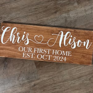 May include: A wooden sign with the text "Chris & Alison Our First Home Est. Oct 2024" in white lettering.