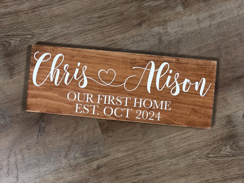 May include: A wooden sign with the text "Chris & Alison Our First Home Est. Oct 2024" in white lettering.