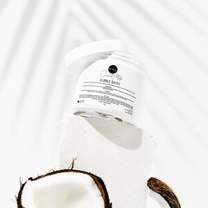 May include: White jar of Coconut Milk Bubble Bath on a white platform with a halved coconut in the foreground. The jar has a black label with the text "Coconut Milk Bubble Bath".