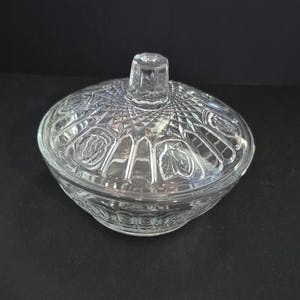 May include: Clear glass candy dish with a lid. The dish has a round shape with a decorative pattern on the lid and the base. The lid has a small, square handle. The glass is transparent, allowing the contents to be visible.