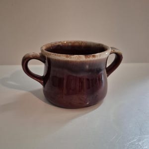 May include: A brown ceramic mug with two handles. The mug has a dark brown base that fades to a lighter brown and cream color at the rim. The mug is designed with two curved handles for easy gripping.