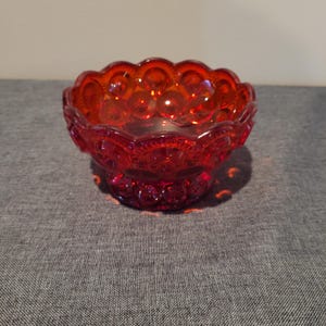 May include: A ruby red glass bowl with a scalloped rim and a star and moon pattern. The bowl sits on a small pedestal base. The glass is translucent, allowing light to pass through, and is likely vintage.