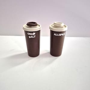 May include: Two brown spice shakers with off-white tops. One shaker is labeled "ONION SALT" and the other "ALLSPICE" in white text. The shakers are cylindrical and sit on a white surface.
