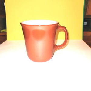 May include: A terracotta colored ceramic mug with a white interior and a rounded handle.