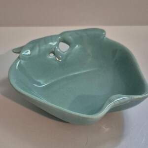 May include: A light blue ceramic dish with a leaf-like design. The dish has a small handle and a hole near the top. The dish is approximately 6 inches long and 5 inches wide.