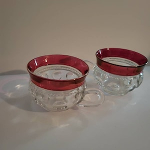 May include: Two clear glass cups with red rims. The cups have a textured pattern and a handle.