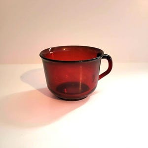 May include: A red glass cup with a handle. The cup is made of clear glass with a red tint.