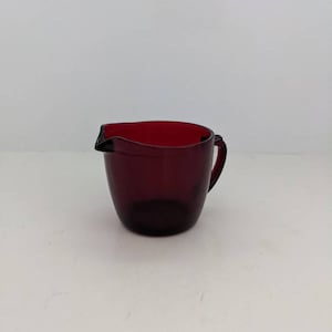 May include: A small, dark red glass creamer with a handle. The creamer is made of glass and has a smooth, rounded shape.