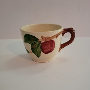 May include: A white ceramic teacup with a brown handle and a painted design of red apples and green leaves on a branch.