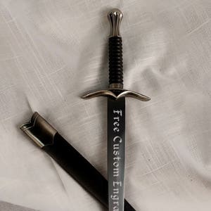 May include: A silver dagger with a black handle and a silver scabbard. The blade is engraved with the words "Free Custom Engraving!"