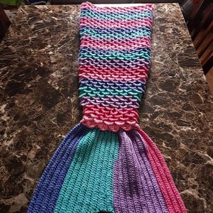 May include: A crocheted mermaid tail blanket in shades of pink, purple, teal, and blue. The tail is made of a textured fabric and has a striped pattern.