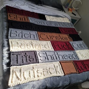 Könnte beinhalten: Eine handgefertigte Häkeldecke mit verschiedenen farbigen rechteckigen Feldern. Jedes Feld zeigt ein anderes Wort oder eine andere Phrase, wie "Arschloch" und "Hure". Die Decke liegt auf einem Bett.
