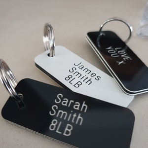May include: Three rectangular keyrings with silver hoops. One is white with "James Smith 8LB" in silver text. Another is black with "Sarah Smith 8LB". The third is black with "LOVE YOU X" in silver text.