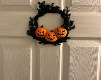 Może przedstawiać: Wieniec na Halloween z trzema pomarańczowymi dyniami z wyrzeźbionymi twarzami, otoczony sylwetkami czarnych kotów i gałęziami. Wieniec jest zamontowany na białych drzwiach, tworząc świąteczną dekorację sezonową.
