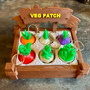 May include: A brown wooden display with the text "VEG PATCH" in yellow, holding six vegetable-shaped keychains. The keychains feature a carrot, turnip, radish, and lettuce, each with a gold keyring. The vegetables are in a white tray.