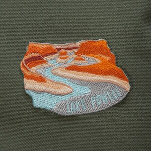 Peut inclure: Un sac à dos en toile vert avec un patch qui dit "Lake Powell" et représente un paysage avec une rivière et des falaises orange.