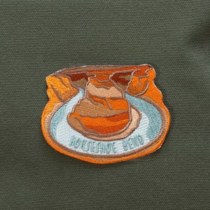 Può includere: Uno zaino in tela verde con una toppa ricamata marrone e arancione che dice "Horseshoe Bend".