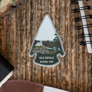May include: Arrowhead-shaped sticker featuring a landscape scene from Isle Royale National Park. The sticker is on a wooden surface, alongside a camera, notebook, and camera strap. The sticker has a white border and the text "Isle Royale National Park".