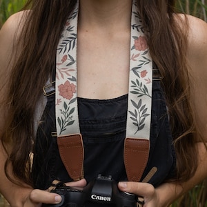Pode incluir: Uma alça de câmera com um design floral de flores rosa e folhas verdes em um fundo creme. A alça tem extremidades de couro marrom. Uma câmera Canon preta é segurada em frente a plantas verdes altas.