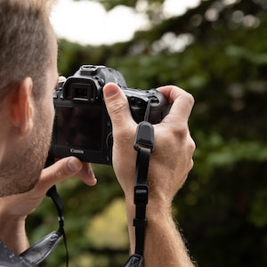 May include: A black Canon digital camera featuring a rear LCD screen, a viewfinder, and a black adjustable camera strap with a plastic buckle, held in a horizontal orientation to capture a photograph of the surrounding outdoor environment.