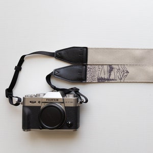 May include: A silver camera with a black leather strap and a beige camera strap with a mountain scene printed on it.