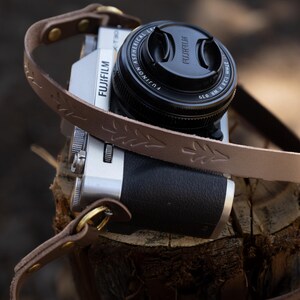 Może przedstawiać: Srebrny aparat Fujifilm z czarną osłoną obiektywu i brązowym skórzanym paskiem z wzorem liści. Aparat spoczywa na pniu drzewa, z paskiem przewieszonym wokół niego. Obiektyw aparatu jest czarny.