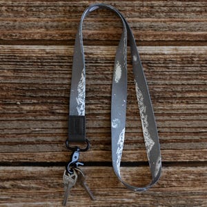 May include: A grey lanyard featuring a mountain pattern, with a black clasp and two keys attached. The lanyard is made of a soft material and is designed to be worn around the neck.
