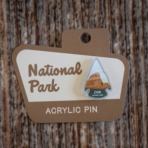 May include: A brown and white acrylic pin with a graphic of a mountain range and the text "Zion National Park". The pin is on a brown card with the text "National Park" and "Acrylic Pin".