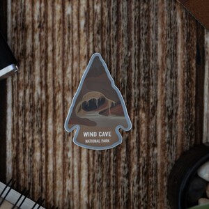 Puede incluir: Una pegatina en forma de flecha marrón y beige con una representación de una cueva y el texto "WIND CAVE NATIONAL PARK".