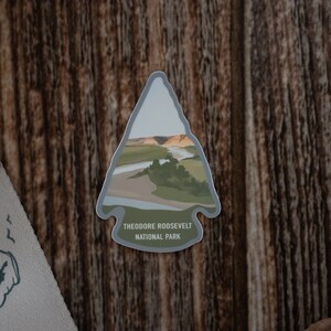Puede incluir: Una pegatina gris en forma de flecha con una ilustración de paisaje de un río y árboles. La pegatina dice "Theodore Roosevelt National Park".