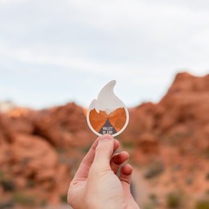 Puede incluir: Una pegatina blanca con un diseño de llama y el texto "Valley of Fire" en negro. La pegatina se está sosteniendo frente a un fondo borroso de formaciones rocosas rojas.