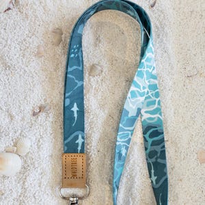 Könnte beinhalten: Ein blaues Schlüsselband mit Hai-Muster und einem Lederanhänger mit der Aufschrift "WALDTREE". Das Schlüsselband ist an einem silbernen Verschluss befestigt, der zwei Schlüssel hält. Der Hintergrund ist weißer Sand mit Muscheln.