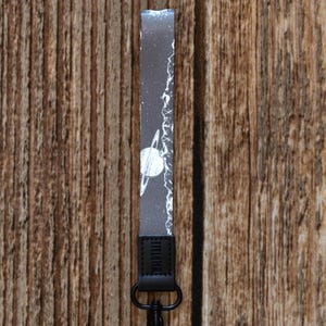 May include: A lanyard with a black leather strap and a black metal clasp holding two keys. The lanyard features a grey and white space-themed design with a planet and stars. The lanyard is hanging against a wooden background.