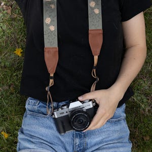 May include: A Fujifilm X-T30 camera with a black body and silver top plate, attached to a sage green camera strap featuring a repeating pattern of small, light-colored roses. The strap includes brown leather end tabs and thin nylon connectors.