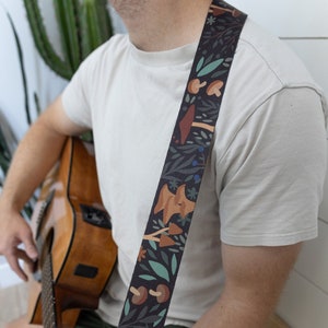 May include: A person playing an acoustic guitar with a black guitar strap featuring a colorful mushroom and floral pattern.