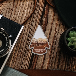 May include: A gray arrow-shaped sticker with a brown and white image of a landscape and the text "Petrified Forest National Park".