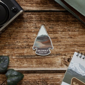 May include: A brown and gray arrow-shaped sticker with a mountain scene and the text "Haleakala National Park".