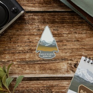 May include: A gray and white arrow-shaped sticker with a mountain scene and the text "Gates of the Arctic National Park".