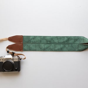 Pode incluir: Uma correia de câmera verde com um padrão de folhas e detalhes em couro marrom. A correia está presa a uma câmera prateada.