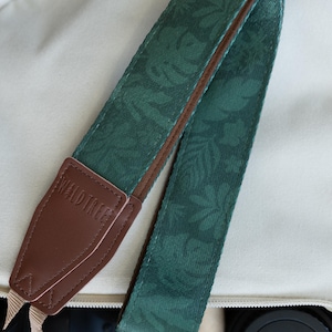 May include: A camera strap featuring a dark green fabric with a subtle leaf pattern. The strap has brown leather end tabs embossed with the text WALDTREE and includes beige nylon webbing connectors attached to a black camera body.
