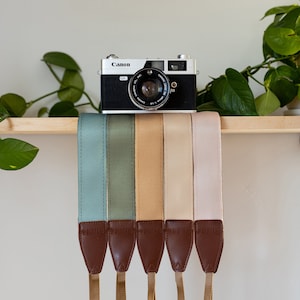 Może przedstawiać: Sześć pasków do aparatu w różnych kolorach, w tym turkusowym, szarozielonym, beżowym i pudrowo różowym, jest wystawionych na drewnianej półce. Paski są wykonane ze skóry i mają brązowy skórzany akcent na dole. Vintage'owy aparat Canon stoi na półce przed paskami.