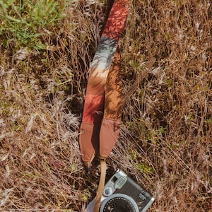 laying in the grass is a retro striped printed camera strap. Striped pattern of shades of red, orange, brown ,blue, and green. Camera connected to film camera.