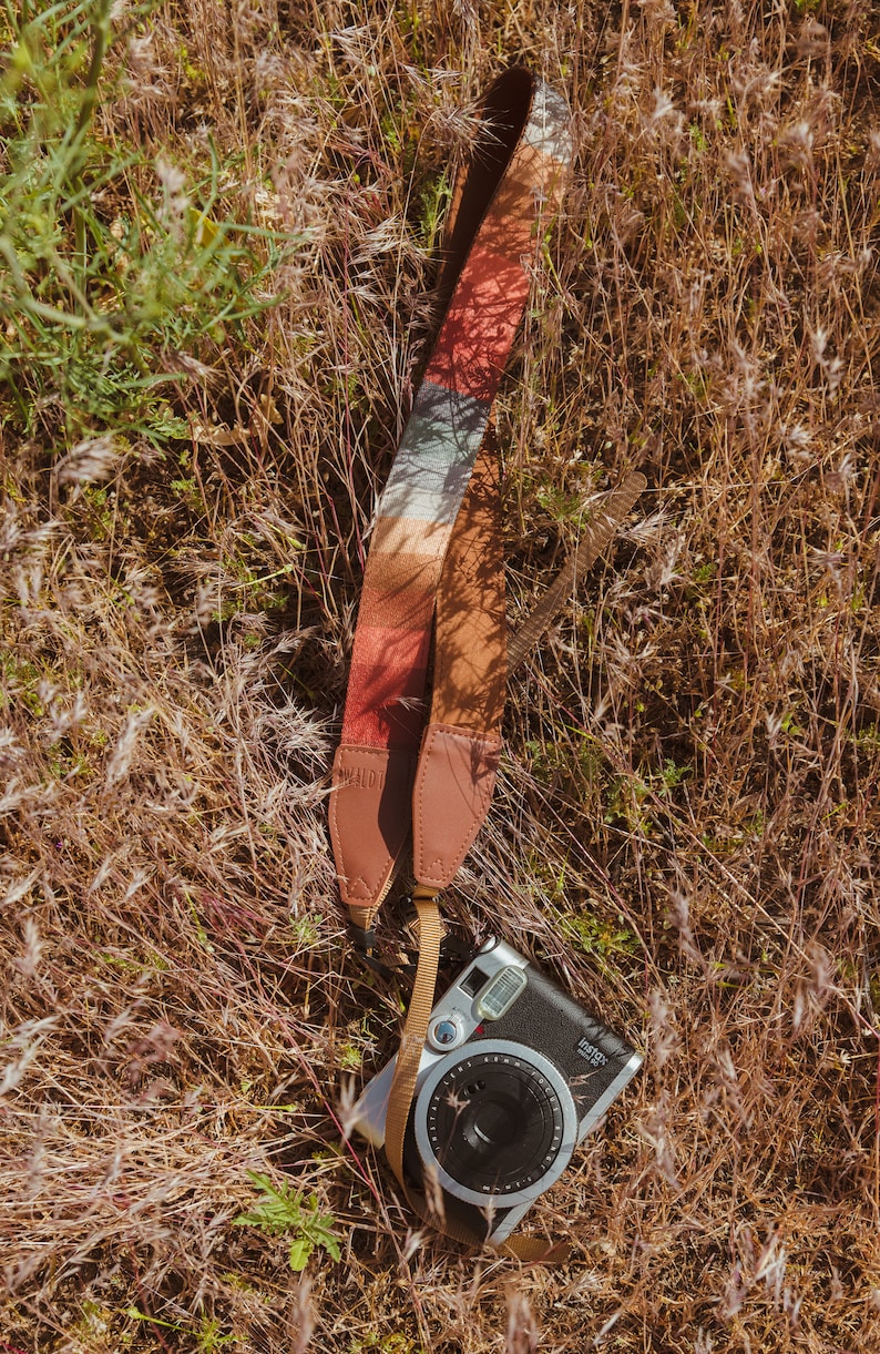 laying in the grass is a retro striped printed camera strap. Striped pattern of shades of red, orange, brown ,blue, and green. Camera connected to film camera.