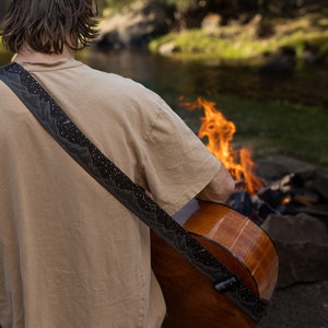 Man sitting in front of campfire playing acoustic guitar wearing a black guitar strap printed in black white white stars and dark gray mountains printed across the length of the strap