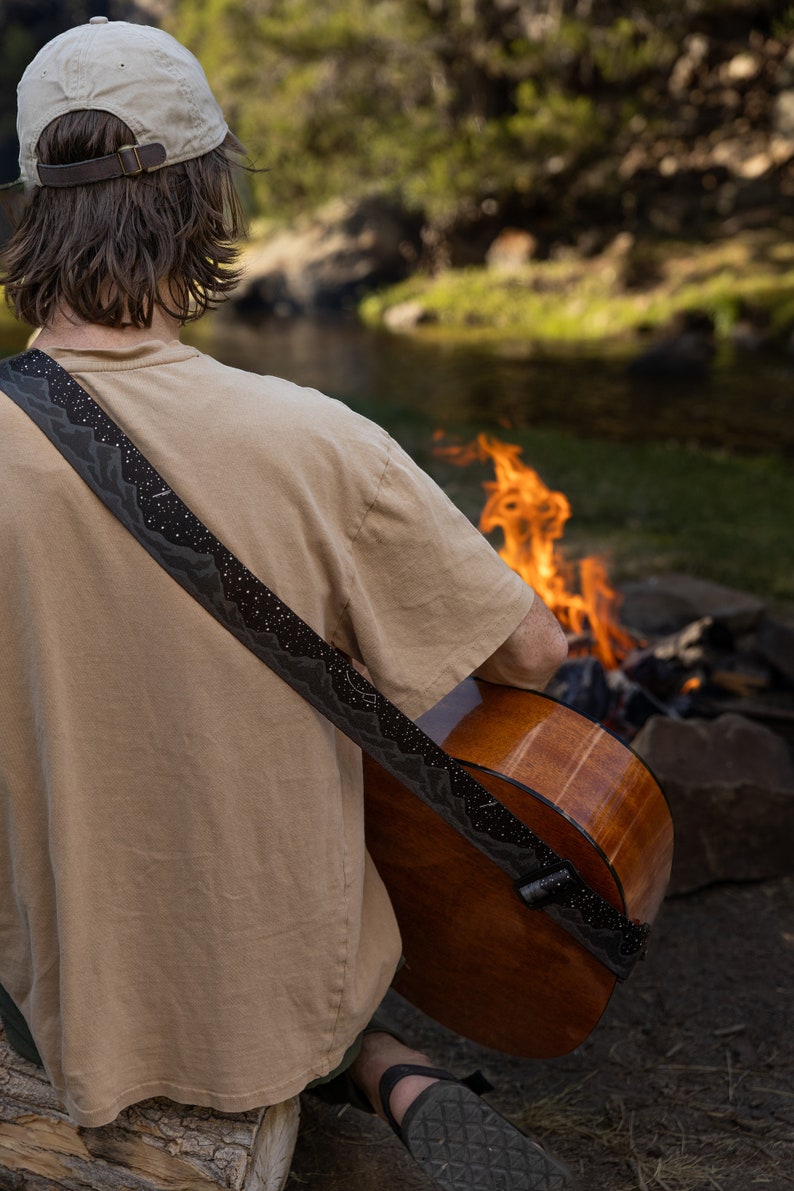 Man sitting in front of campfire playing acoustic guitar wearing a black guitar strap printed in black white white stars and dark gray mountains printed across the length of the strap