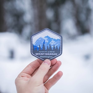 May include: A blue and white sticker with a mountain range illustration and the text "9,131 FT 2,783 M MOUNT SHUKSAN Washington".