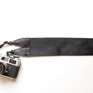 May include: A silver Canon camera with a black leather strap. The camera has a black lens and a black body.