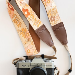 May include: A camera strap featuring a floral design in peach, orange, and yellow on a cream backdrop. The strap has brown leather ends with the text "WALD TREE". A Fujifilm X-T30 camera with a black lens cap is below.