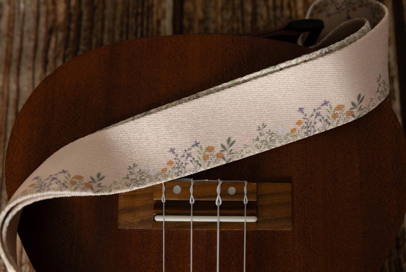 Cute tan floral strap printed with Little colored flowers across the length of the strap attached to Ukulele lying on wood floor