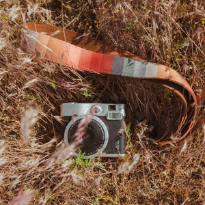laying in the grass is a retro striped printed camera strap. Striped pattern of shades of red, orange, brown ,blue, and green. Camera connected to film camera.